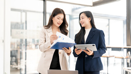 Two professional Asian businesswomen discussing financial reports and data on a tablet in a bright, modern office. Teamwork and success concept.