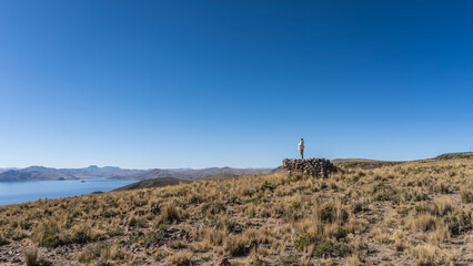 The high mountain region of the Andes. Dry yellowed grass on the plateau. A silhouette of a man is visible on a stone pedestal, looking into the distance. Beautiful lake, mountain range on the horizon
