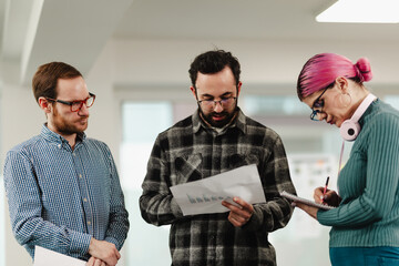 Three colleagues collaborate in a modern office, reviewing documents and charts, taking notes during a casual team meeting as they discuss data, ideas, and project progress