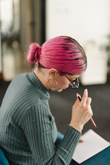 Businessperson with pink hair engaged in note-taking at a modern office setting during a productive meeting