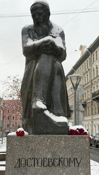 A snow-covered statue of a seated figure, possibly a writer, with flowers at the base, set in an urban environment