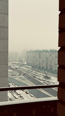 A snowy urban landscape viewed from a balcony, featuring tall apartment buildings and a road with parked cars, under a gray sky