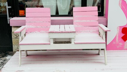 A pink and white outdoor bench covered in snow, situated in front of a shop window
