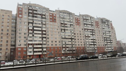 A row of modern apartment buildings in a snowy urban setting, with parked cars in front and a cloudy sky