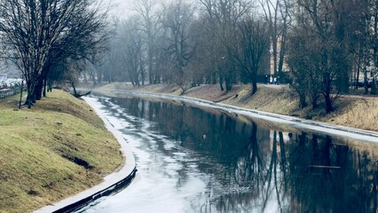 A tranquil river scene with calm water reflecting trees and a cloudy sky, surrounded by grassy banks, creating a serene atmosphere