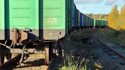 A close-up view of a green freight train car on tracks, with a long line of train cars curving in the background, surrounded by autumn foliage