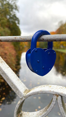 A blue heart-shaped padlock attached to a metal railing on a bridge, with a river and autumn trees in the background.