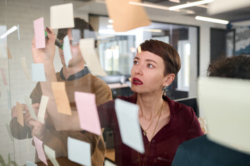 Caucasian young adult woman brainstorming ideas with colleagues, writing on transparent board covered with sticky notes, collaborating in modern office environment during meeting