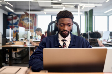 Young adult Black man sitting at desk working on laptop in modern open plan office, focused expression, other people collaborating in background, glass partition visible