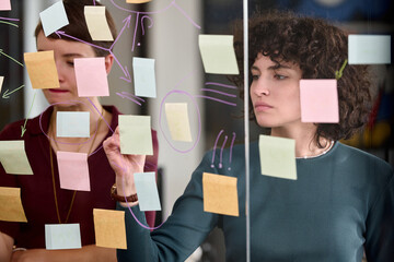 Young adult Caucasian women collaborating on project, writing and organizing sticky notes on glass wall, focusing on brainstorming and planning tasks