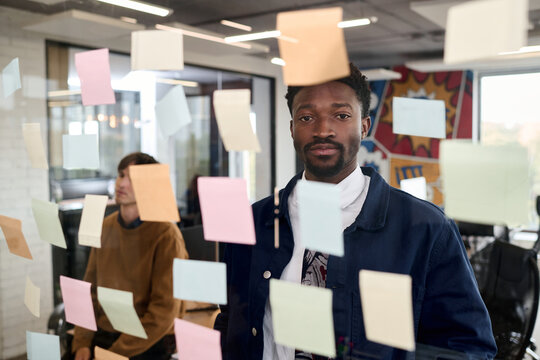 Portrait of young adult Black man standing in modern office looking into camera surrounded by colorful sticky notes on glass wall with Caucasian young adult man working in background