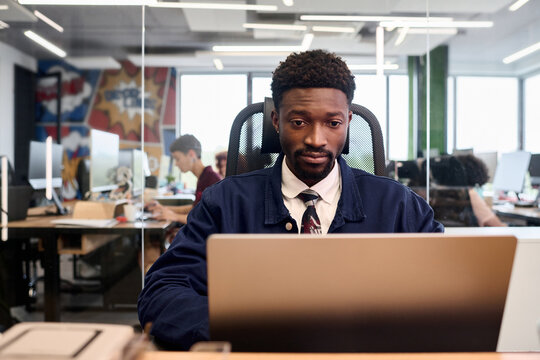 Young adult Black man sitting at desk working on laptop in modern open plan office, focused expression, other people collaborating in background, glass partition visible