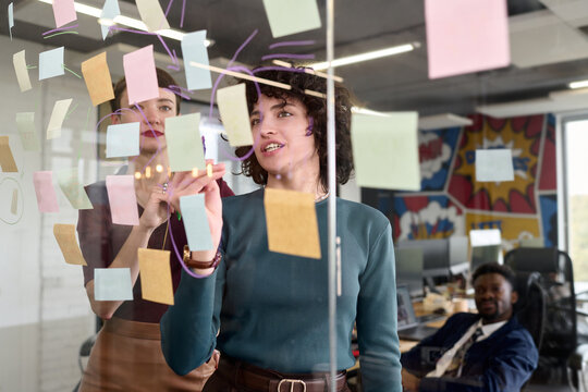 Young adult Caucasian woman and young adult Black woman collaborating and brainstorming ideas on transparent board with sticky notes in modern office while young adult Black man observing