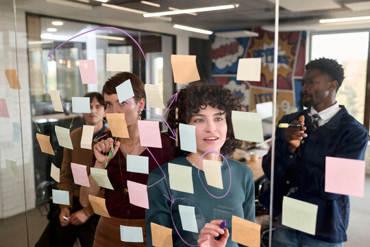 Multiethnic group of young adult women and man brainstorming and drawing diagrams on glass wall covered with sticky notes in modern office, collaborating on creative project