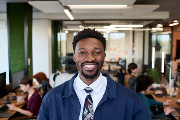 Portrait of young adult Black man smiling confidently while standing in modern office workspace, open plan environment with diverse coworkers working at desks in background