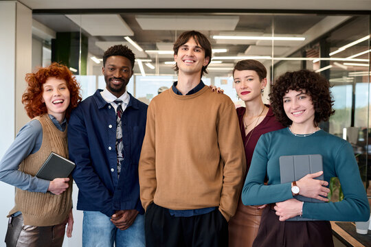 Group of diverse young adults standing together in modern office, smiling and looking at camera, including Caucasian women, Black man, Caucasian man, holding digital tablets