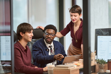 Young adult Caucasian man, young adult Black man, young adult Caucasian woman collaborating in modern office, discussing project at desk with laptop and documents, working together