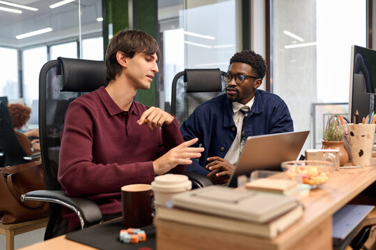 Caucasian young adult man explaining project details to Black young adult man while sitting at desk with laptop in modern office, both focused on business discussion