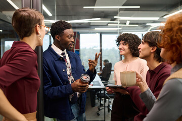 Group of young adult multiethnic colleagues standing in modern office, engaging in lively conversation, Black man gesturing while holding reusable coffee cup, others listening and smiling