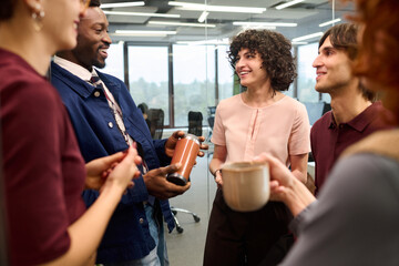 Group of diverse young adults standing in modern office, smiling and talking during coffee break, multiethnic colleagues interacting and holding mugs, casual business meeting atmosphere