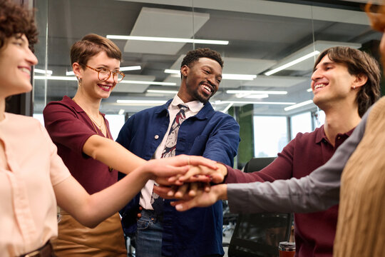 Diverse group of young adults standing in modern office joining hands in center smiling and interacting with each other showing teamwork and collaboration during meeting