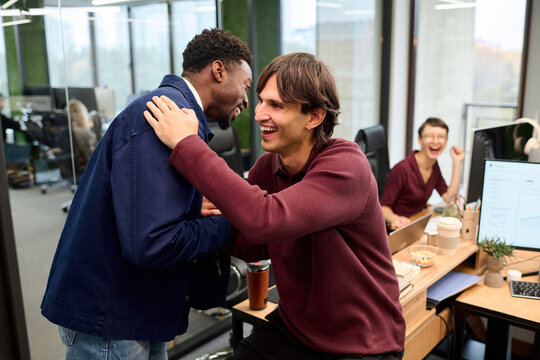 Caucasian young adult man greeting Black young adult man with handshake and hug in modern office while smiling woman sitting at desk in background laughing and watching interaction