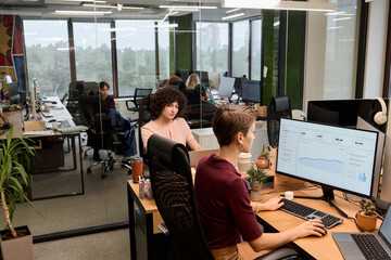 Young adult Caucasian women working at desks in modern open plan office, focusing on computer monitors displaying analytics charts, colleagues visible in background