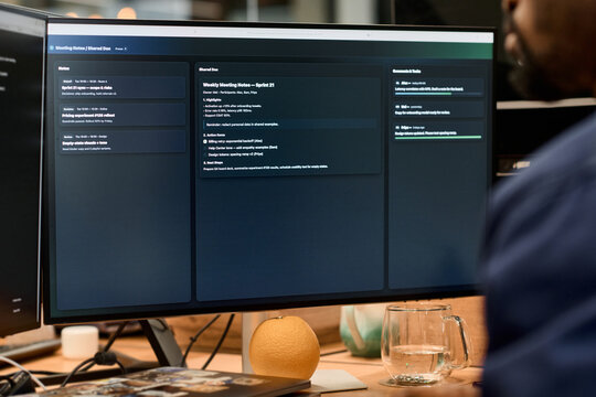Black man sitting at desk working on computer monitor displaying project management dashboard, glass of water and orange on wooden surface, office environment visible in background