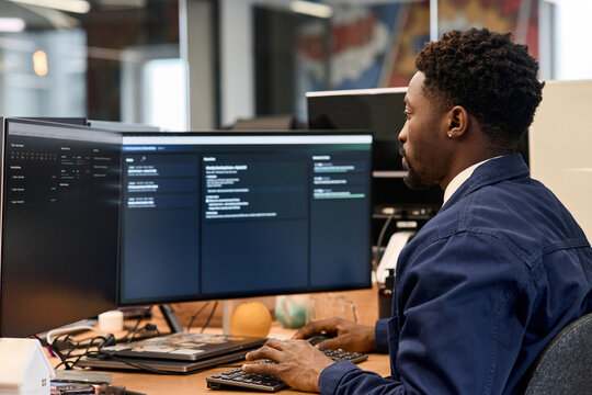 Young adult Black man working at desk using dual monitors in modern office, focusing on computer screen while typing on keyboard, short hair visible, professional environment