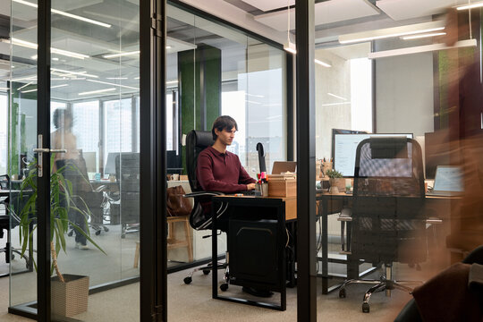 Young adult Caucasian man sitting at desk working on computer in modern office, focused expression, glass walls and open workspace visible, colleagues moving in background
