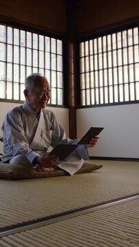 Elderly japanese man sitting on a tatami mat in traditional wear, reading a book inside a room with shoji screens letting in natural light during a serene moment
