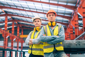 Portrait confident Caucasian industrial engineer men arms crossed standing on factory catwalk. Professional male worker partner wear safety helmet vest earmuffs. Heavy metal manufacturing team leader