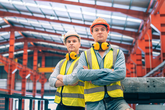 Portrait confident Caucasian industrial engineer men arms crossed standing on factory catwalk. Professional male worker partner wear safety helmet vest earmuffs. Heavy metal manufacturing team leader