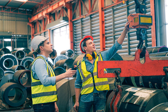 Caucasian industrial engineer men controlling overhead crane with remote in factory. Worker partner wear safety helmet vest lifting heavy metal coil material. Manufacturing hoist machine logistics