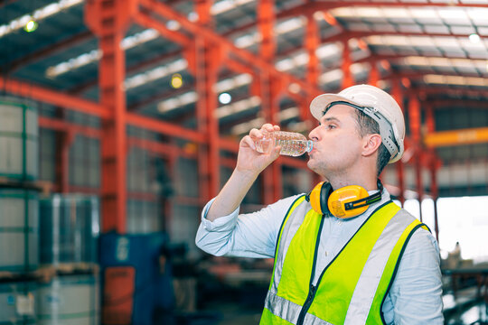 Thirsty Caucasian industrial engineer man wear safety helmet vest drinking water from plastic bottle in factory. Tired worker taking break refreshment. Manufacturing plant hydration health care