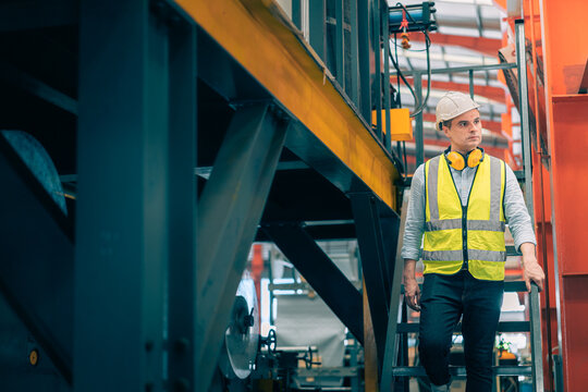 Caucasian industrial engineer man wear safety helmet vest walking down metal stairs in factory. Professional worker holding handrail safety routine. Heavy manufacturing plant background.