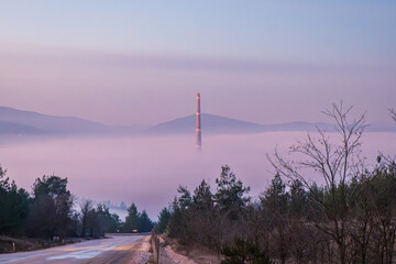 Tall industrial chimney stack rising above a thick layer of pink fog in a valley with a road in the foreground at purple sunrise. Orhaneli, Bursa, Turkey.