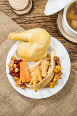 A plate of boiled chicken served with roots, berries, and herbs beside a ceramic soup pot.