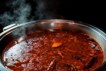A stainless steel pot filled with rich red chili broth and floating herbs served on a wooden table.