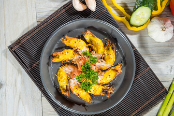A round black plate with grilled shrimp arranged in a circle, garnished with parsley, surrounded by fresh vegetables on a wooden mat.