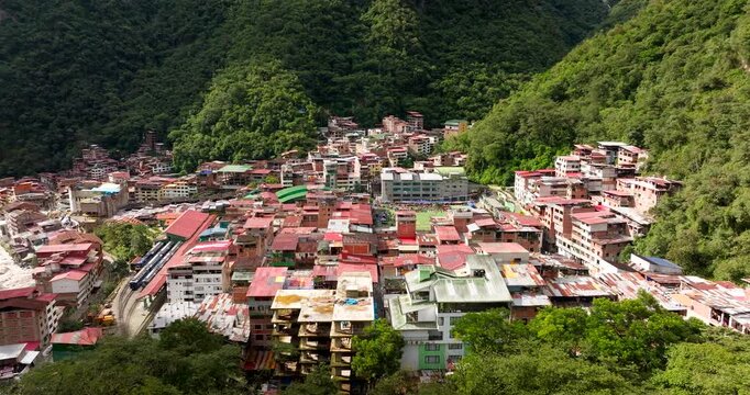 Cinematic aerial drone view of Aguas Calientes, the gateway town to Machu Picchu. The Urubamba River flows alongside the colorful buildings nestled in the green Andes mountains. Ideal for Peru travel