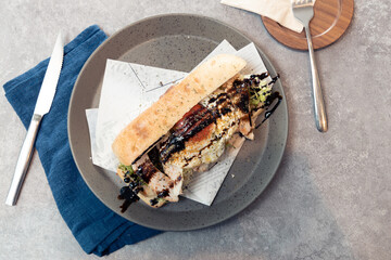 A long sandwich filled with grilled chicken, vegetables, and balsamic glaze served on a gray plate with a knife and fork on the side.
