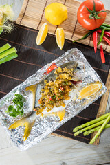 A whole fish grilled and topped with savory sauce, served on foil with lemon wedges and fresh vegetables for garnish on a wooden mat.