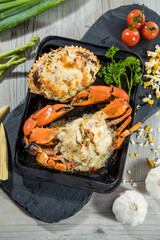 A black tray with two crabs topped with melted cheese and herbs, surrounded by fresh vegetables and garlic for a rich seafood presentation.