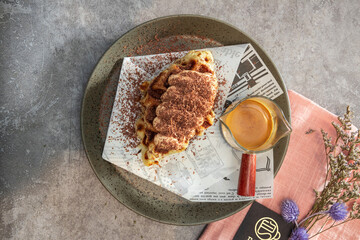 A plate of tiramisu topped with cocoa powder served on printed paper, accompanied by a glass of espresso and cinnamon stick on a gray surface.