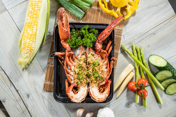 A black tray with a halved lobster topped with garlic and herbs, surrounded by fresh vegetables including corn, tomato, and asparagus.