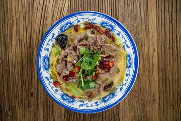 A blue floral bowl filled with thin beef slices in sour broth, garnished with chili peppers and coriander, served on a wooden table.