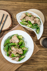 Two plates of stir-fried pork slices with fresh green okra and garlic, served on a wooden table with chopsticks for a rustic presentation.