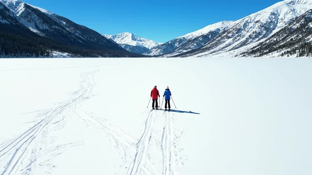 Aerial view of couple cross-country skiing on frozen lake towards mountains. Sunny winter day representing active lifestyle and freedom. Perfect for travel or sport concepts.