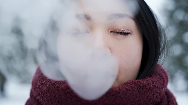 Close up of young Asian woman with frost on eyelashes exhaling visible breath in cold winter forest. Atmospheric portrait for winter holidays, skincare, and seasonal concepts.